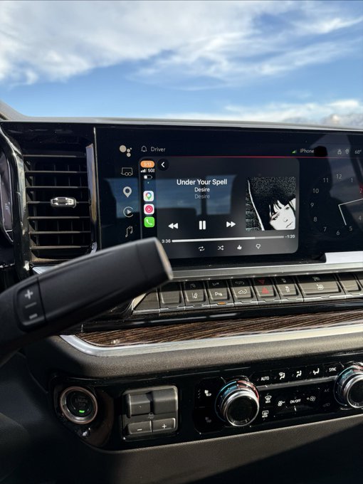 Interior dashboard view of a vehicle with infotainment screen displaying the song 'Under Your Spell' by Desire playing via CarPlay.
