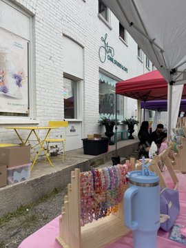 The image shows an outdoor market setup in front of a storefront, with a table displaying colorful handmade accessories like beaded bracelets and scrunchies on display stands, accompanied by a red Coca-Cola umbrella and yellow bistro tables. The caption references "The Old Exchange" sign on the building, aligning with the post about supporting a small business at the Kitchener, ON market.