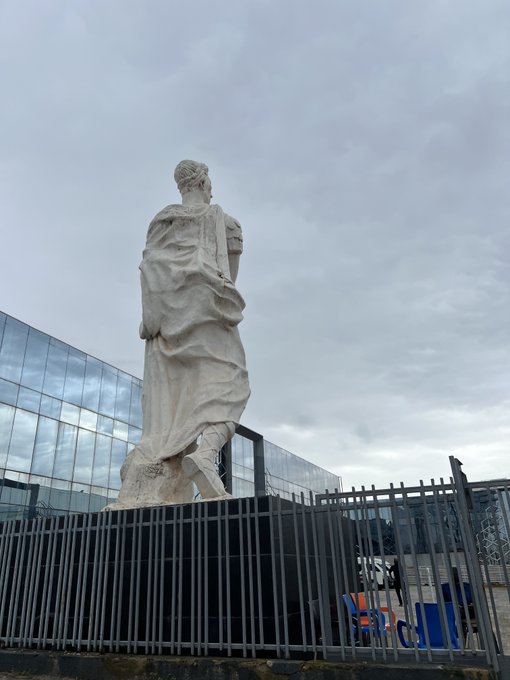 A large white classical statue of a robed figure stands behind a metal fence with a modern glass building and colorful chairs visible in the background under a cloudy sky.