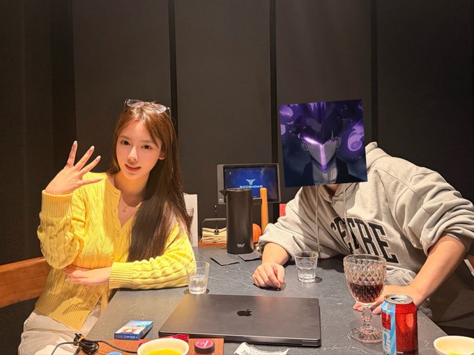 Two people sitting at a table with laptops, drinks, and a red Coca-Cola can; one person's face is covered with an image of a purple mecha/robot character.
