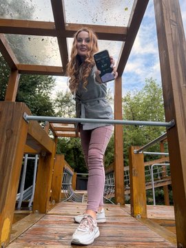 A woman stands on a wooden outdoor structure holding up a smartphone, displaying what appears to be the StepN app on the screen.