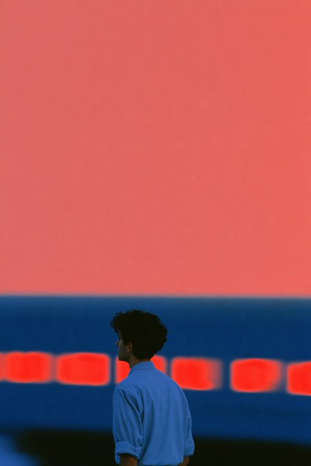 A stylized photograph of a young man in a blue shirt standing in profile against a striking red and blue background with out-of-focus lights.