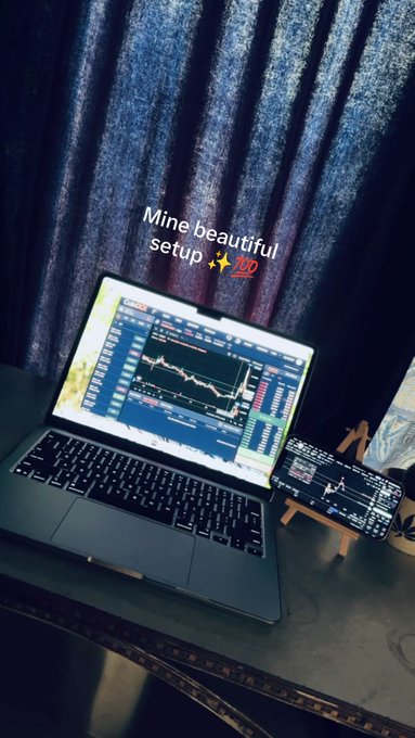 A laptop and phone display trading charts with the caption 'Mine beautiful setup ✨💯' against a dark curtain backdrop.