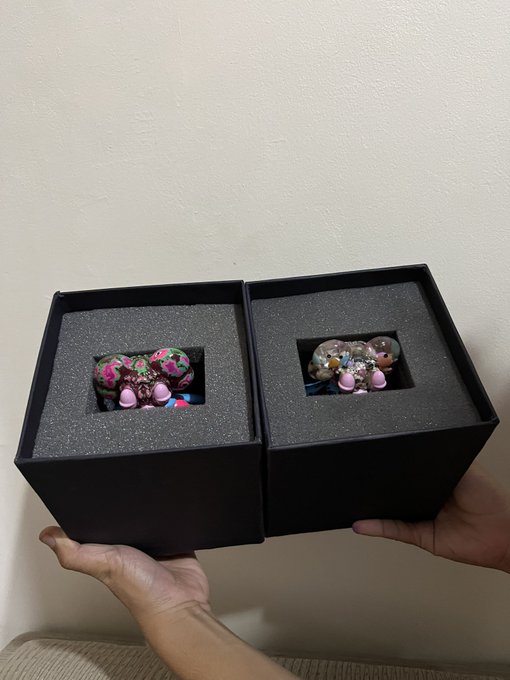 Two black boxes with foam inserts containing small decorative figurines, one featuring pink and green designs and the other with shiny bead-like details.