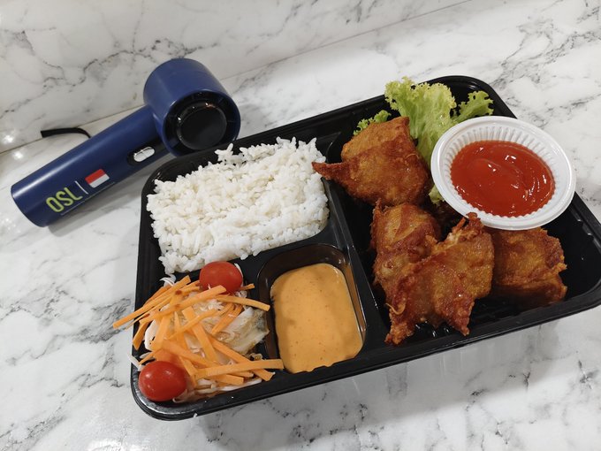 A meal prep container on a marble surface with white rice, fried chicken, lettuce, a small salad, and two dipping sauces, alongside a navy hair dryer branded 'OSL'.