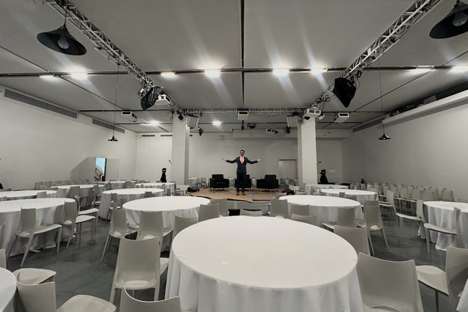 A large event hall with round white tables and chairs set up around a central stage where a person stands with arms outstretched under theatrical lighting.
