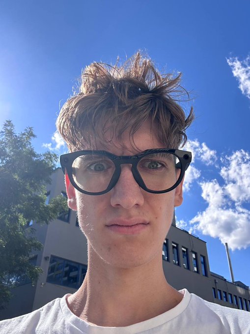 A close-up selfie of a young man with tousled brown hair wearing black-rimmed glasses and a white shirt, photographed from a low angle against a blue sky with clouds and a building in the background.