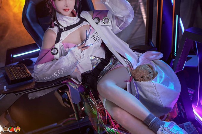 A cosplayer in an elaborate white and purple futuristic outfit sits in a gaming chair holding a small teddy bear, with neon lighting in the background.
