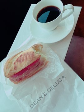 A photograph of a coffee cup on a saucer and a sandwich wrapped in paper with 'DEAN & DELUCA' printed on it, served on a wooden table.