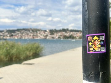 A square sticker on a black pole featuring a character design and a QR code, with a body of water, a paved walkway, and a hillside town in the blurred background.