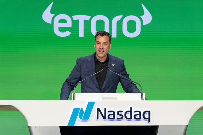 A man in a navy blue suit speaks at a Nasdaq podium, with a large green backdrop displaying the 'eToro' logo with horns, likely from the company's IPO listing ceremony.