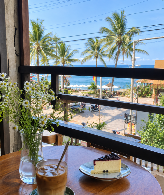 A scenic café view from a balcony overlooking a tropical beach with palm trees, featuring a table set with an iced coffee, a slice of blueberry cheesecake, and a vase of daisies.