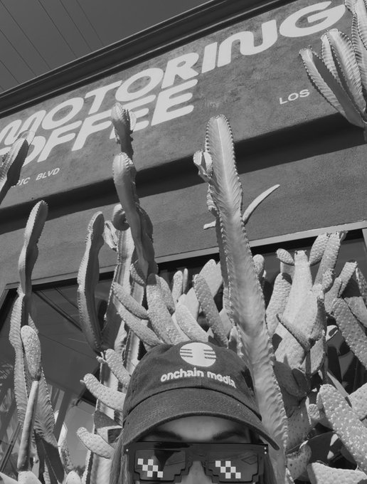 A person wearing an "onchain media" cap and pixelated sunglasses poses in front of a "MOTORING COFFEE" sign, with cacti visible behind them. The black-and-white image captures the LA vibe of the "Cars, Crypto & Coffee" event mentioned in the post.