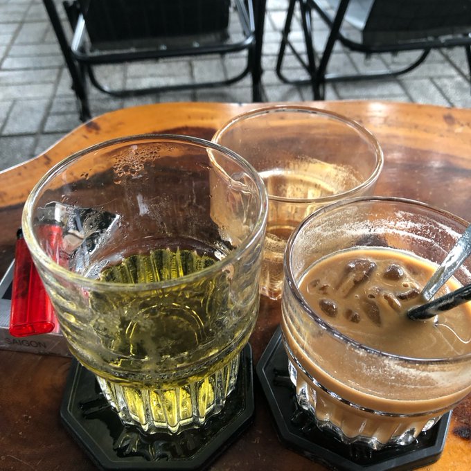 Three glasses sit on a wooden table at a café, including a glass of yellowish liquid, a clear drink, and an iced coffee with milk, alongside a red lighter on a coaster.