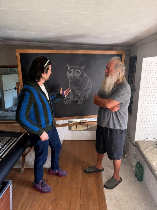 Two men stand on either side of a large chalkboard featuring a detailed chalk drawing of a sitting cat, with one gesturing toward the artwork while the other listens with arms crossed. The casual studio setting and focus on traditional chalk drawing technique captures the essence of "back to basics" artistic practice.