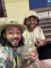 Two men wearing hats smile at the camera at what appears to be a convention, with one making a peace sign.