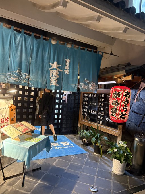 A person enters a Japanese restaurant adorned with teal "大江戸温泉" (Oedo Onsen) noren curtains and a red "おしるこ からすみ" lantern, with a welcome mat reading "KAPILAWAN." The image captures the venue's traditional ambiance, setting the stage for the diverse virtual currency meetup described in the accompanying post.