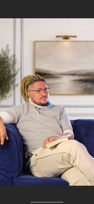 The image features a man sitting on a blue couch with his legs crossed, wearing glasses and a gray shirt. He has dreadlocks and is holding a book while looking off into the distance. The background includes a painting of water and mountains, suggesting that he might be in an art gallery or a room with a scenic view. The caption to the image reads "Them: Litecoin is a dead Dino coin My honest reaction:".