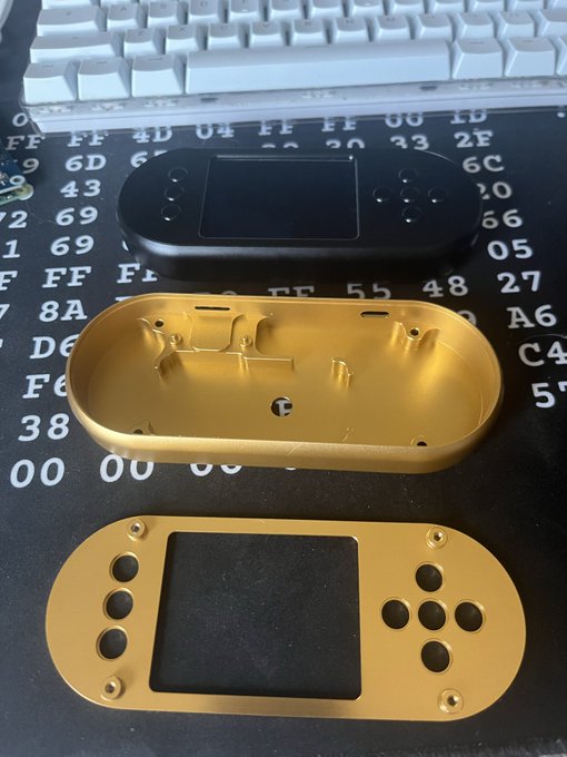 A disassembled handheld game console in matte black and gold casing rests on a desk mat patterned with hex codes like "FF", "6D", "43", and "A6", with a white keyboard in the background—fitting the DIY "Jades" form factor theme as its modular shell is shown in its component parts.