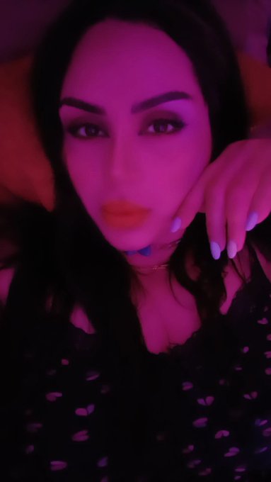 A selfie of a woman with dark hair and heavy eye makeup, wearing a black lace top, illuminated by pink/purple lighting.