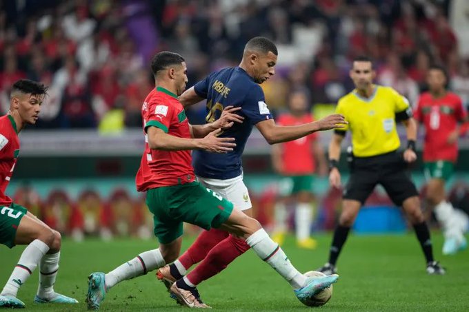 A soccer match showing players from Morocco and Portugal competing for the ball, with Mbappe visible in a France jersey and a referee in yellow.