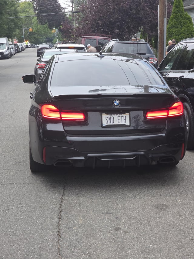 A black BMW with a New York license plate reading "SND ETH" is stopped in traffic on a residential street, playfully personalizing the "Send Ethereum" slang with its custom plate.