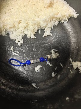 A close-up photograph of a small beaded hair tie with blue and clear beads resting on a dark surface beside a pile of white rice.