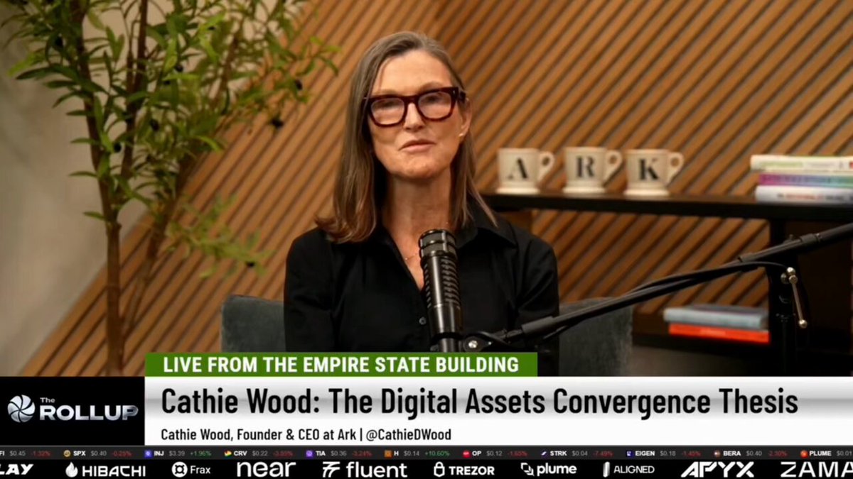 The image shows a woman sitting in front of a microphone, wearing glasses and holding a microphone. She appears to be speaking into it during an interview. The background features a wooden wall with various books arranged against it. The text "Live from the Empire State Building" is visible on the image as well.