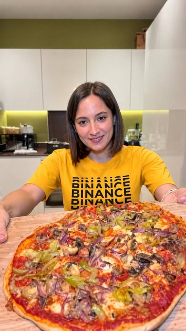 The image features a woman wearing a yellow shirt that says "BinancePizza" as she holds up a large pizza with various toppings such as mushrooms, onions, peppers, and sausage. The background shows a kitchen area with white cabinets and a countertop.