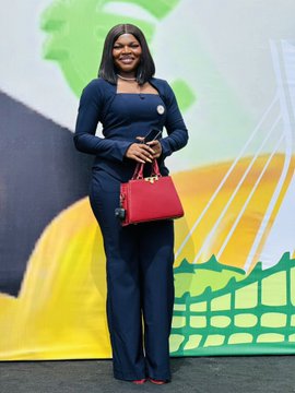 A smiling woman in a navy blue pantsuit stands at what appears to be an event, holding a red handbag and wearing a small name badge, with a colorful map-themed backdrop featuring green and yellow graphics behind her. The professional setting and her polished attire suggest she is attending or speaking at an advocacy event aligned with the "#UnchainSummer26" campaign, though no specific text or names are legible in the image.