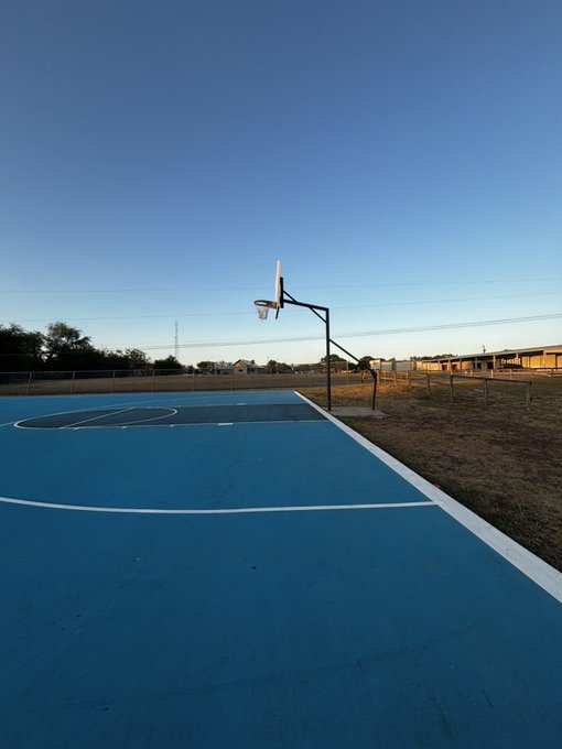 An empty outdoor blue and gray basketball court under a clear sky, with a single hoop and chain-link fencing bordering the playing area.