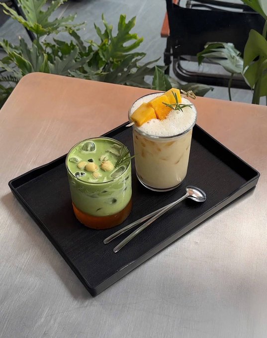 A photo of two iced drinks on a black tray: a green matcha-style drink with ice and a creamy yellow drink topped with mango chunks and rosemary, with a spoon on the side.