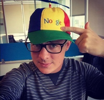 A young man wearing black glasses and a striped shirt points to a multicolored Google-branded trucker hat on his head.