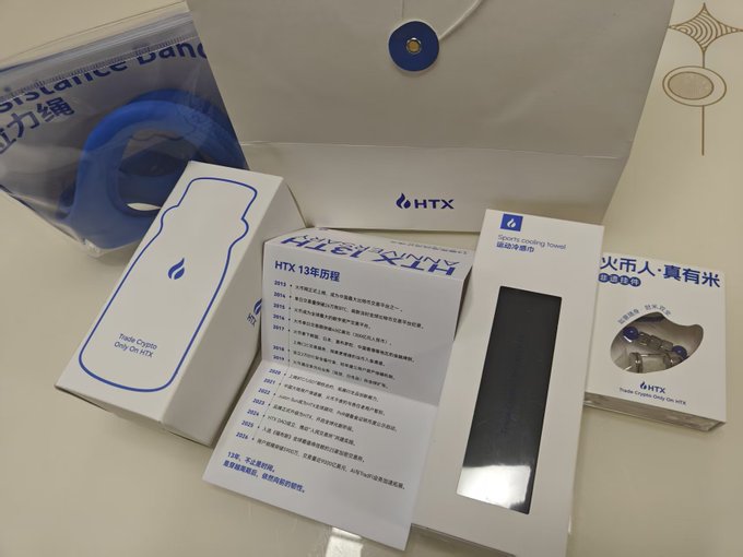 An unboxing photo of HTX (Huobi) promotional merchandise including a Resist silicone band, a paper card listing the company's history, and a box for a sports cooling towel featuring Chinese text.