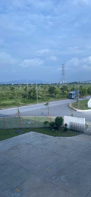 An outdoor scene showing a road with a blue truck passing by, green fields, power transmission towers, and a cloudy blue sky, viewed from a paved area.