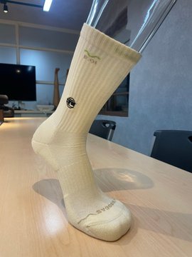 A photograph of a beige crew sock displayed upright on a wooden table, featuring a small black logo on the side and text near the toe.