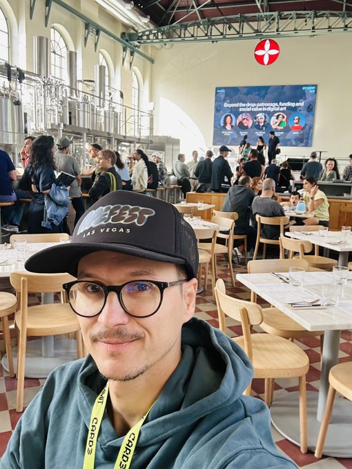 The image features a man wearing glasses, sitting at a table with chairs around him. He has a hat on his head that says "@NFC summit" on it. The man appears to be taking a selfie or capturing a moment from an event he attended. There are several other people in the background who seem to be engaged in conversations and enjoying themselves. A TV screen can also be seen, possibly displaying information about the event or showing some content related to the gathering.