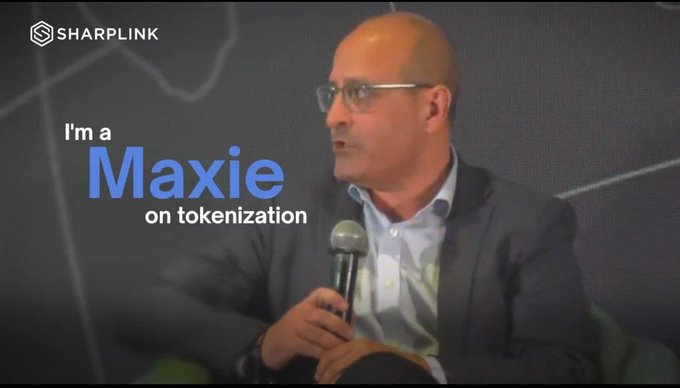 The image features a man wearing glasses sitting down with his hands on his lap while holding a microphone. He appears to be talking or giving a speech at an event, possibly related to cryptocurrency or digital assets. The background of the image shows a blackboard, which could potentially be used for writing or drawing during the presentation. The caption below the image reads: "I'm a Maxie on tokenization".
