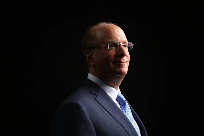 A professional portrait of a middle-aged man with glasses, wearing a dark suit and blue tie, photographed against a black background with dramatic side lighting.
