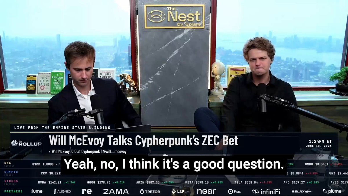 The image shows two men sitting at a desk, with one man wearing a suit and the other wearing a black shirt. They are both talking into microphones, suggesting that they might be participating in an interview or discussion about a topic related to cypherpunk technology. The background of the image features a window overlooking a cityscape, which adds depth to the scene.