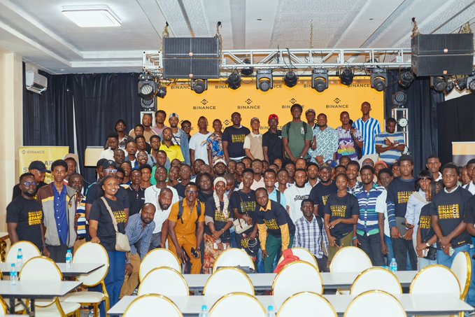 A large group of 196+ attendees pose together at the Binance Freetown Traders Bootcamp in Sierra Leone, with the "BINANCE" branded backdrop visible behind them as they gather in the event hall filled with empty chairs.