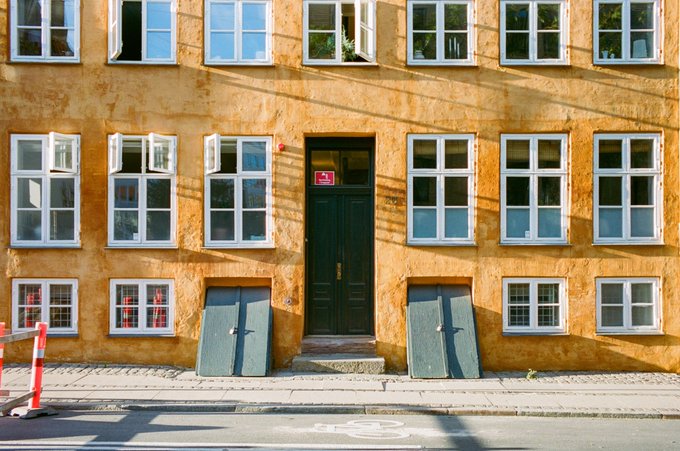 A weathered ochre-colored building facade with numerous white-framed windows and a dark green central door flanked by two closed cellar doors, casting long shadows in the warm sunlight. The traditional architecture, with its symmetrical window arrangement and historic Copenhagen-style design, reflects the cultural and artistic heritage that collectors like @osseneth support through their patronage of community artists.