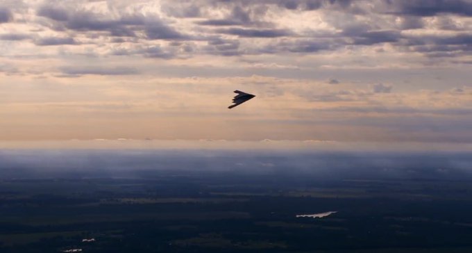 A delta-wing stealth aircraft soars through a dramatic, cloud-filled sky above a hazy landscape, with what appears to be another aircraft or vapor trail visible in the distance below. The futuristic, otherworldly imagery of the advanced jet mirrors the post's reference to "Elysium," evoking a sense of transcending earthly limits toward an idyllic, near-paradise realm.