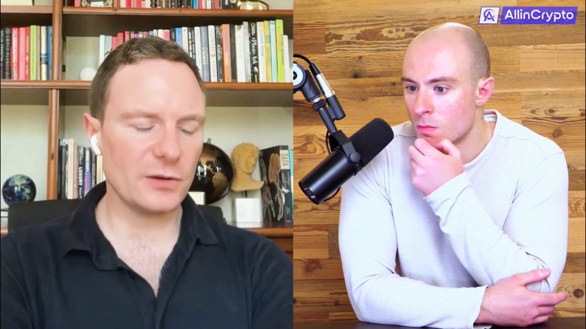 The image shows two men sitting next to each other with their hands resting on a table. One man has short hair and is wearing a black shirt, while the other man has long hair and is wearing a white shirt. They are both looking at something off-camera in front of them. The background features a wooden wall with several bookshelves filled with various books.