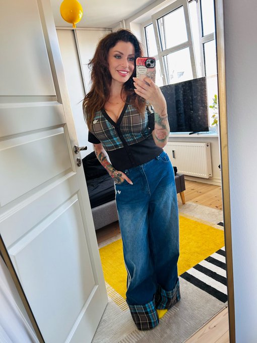 A mirror selfie of a smiling woman with tattoos wearing a plaid top and wide-leg jeans, standing in a room with a yellow balloon and a black-and-white striped rug.