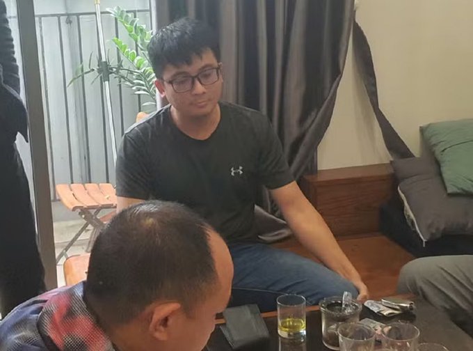 A photo of a young man wearing glasses and a black Under Armour t-shirt sitting on a couch, with another person and drinks visible in the foreground.