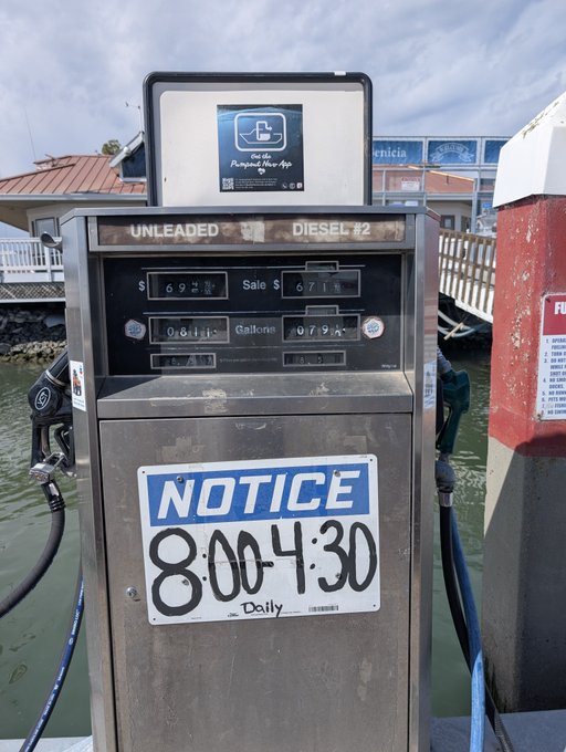 A marine fuel pump displaying prices for 'UNLEADED' and 'DIESEL #2' stands on a dock, with a handwritten sign reading 'NOTICE 8:00-4:30 Daily' and a digital sign prompting to 'Get the Pump & Save App'.