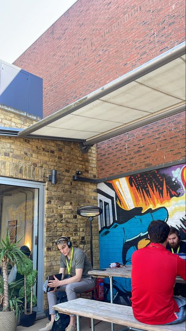 A photograph of an outdoor cafe area with brick walls and a graffiti mural, where a person in a red shirt and another with headphones sit at a wooden table.