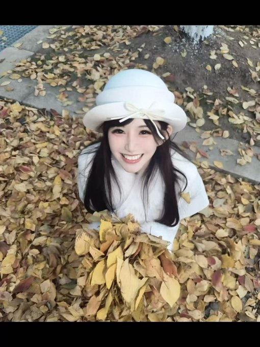A smiling young woman wearing a white bucket hat and cream sweater sits amid a blanket of golden autumn leaves, holding a bundle of them as she looks up at the camera — a cheerful, photogenic autumn portrait that fits the playful "does this count as the best-looking?" vibe perfectly.