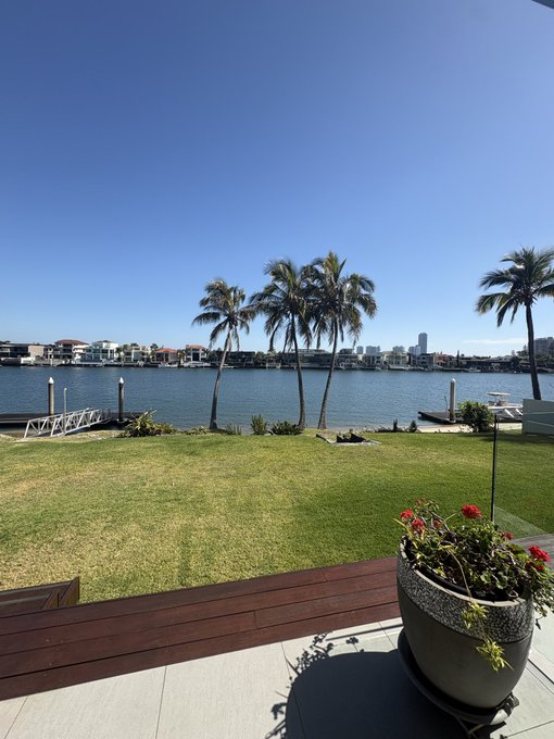A serene waterfront view showcases a lush green lawn framed by swaying palm trees, a calm river, and a distant city skyline under a clear blue sky, with a vibrant potted plant adding a pop of color on the wooden deck—capturing the upscale, family-friendly atmosphere of house hunting in a scenic, affluent neighborhood.

The image complements the post's theme of enjoying life's precious moments with family, as the picturesque setting reflects the luxury home being viewed with the brother-in-law, paired with the cherished time ahead at his son's 10th birthday party.